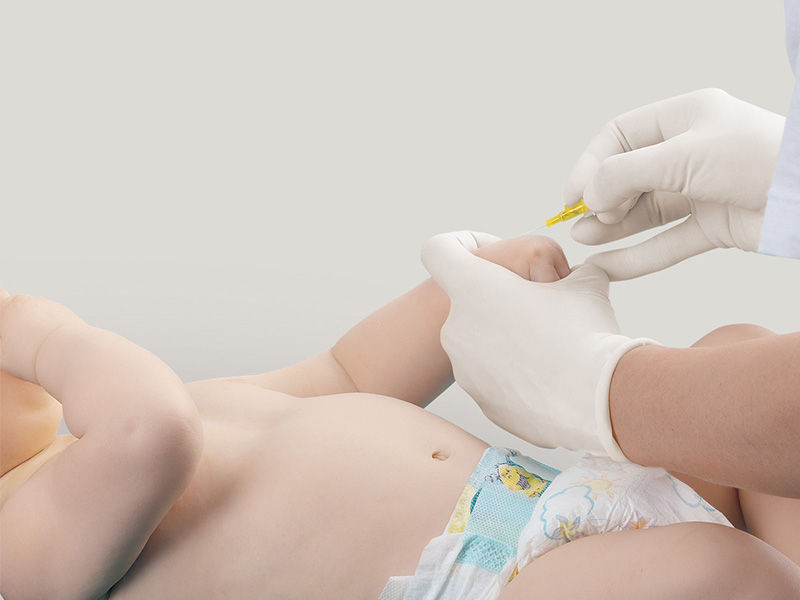Lying baby in diapers. Left arm is held at the wrist by a gloved hand. A gloved right hand holds a yellow Introcan Safety® cannula. The tip of the cannula is pointing towards the puncture site.