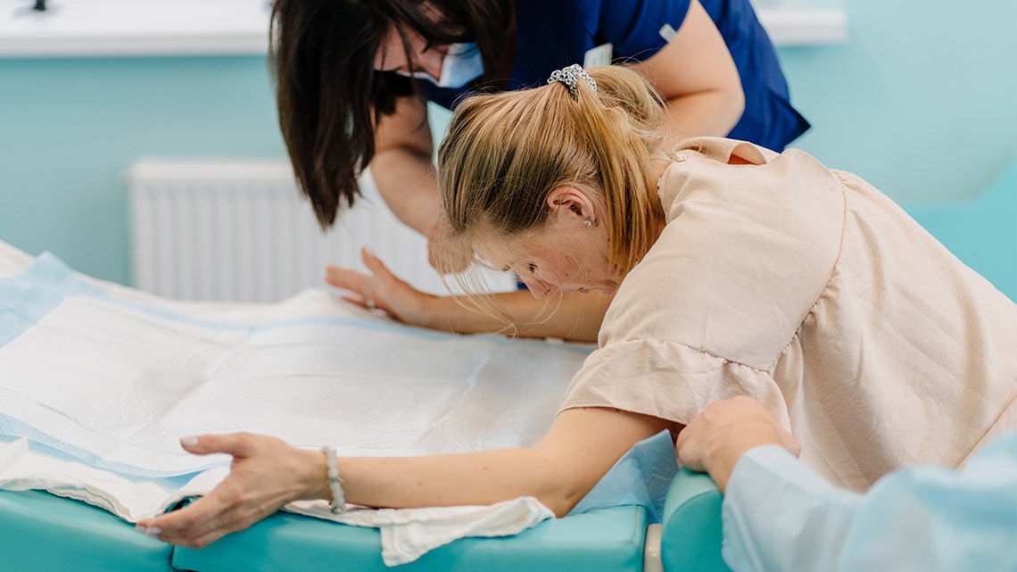 A woman in a healthcare setting, leaning forward on a padded, disposable-covered surface with her arms extended for support. A caregiver in a dark top and mask assists nearby, while another person’s arm offers additional support. The clinical indoor environment features a light wall and radiator, suggesting a moment of physical exertion, likely during childbirth.