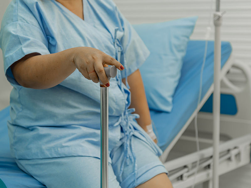 Obese middle-aged female patient in blue hospital clothes sitting on the edge of a bed (blue bed cover). Right hand holds a walking stick. There is a white plaster on her left forearm. An infusion stands next to the bed.