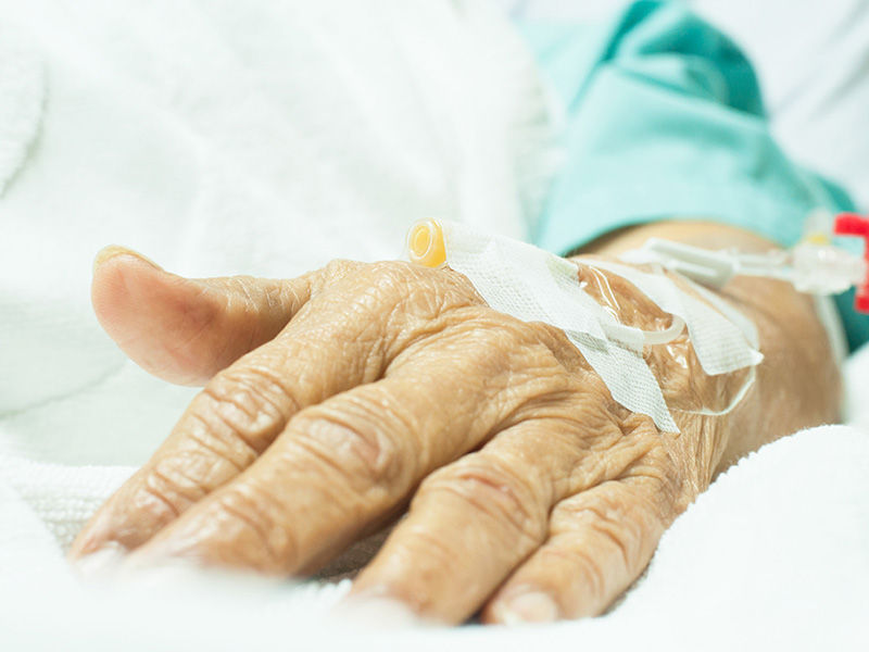 Geriatric, wrinkled hand lying on white bedspread. Puncture site on back of hand. Connected line with white sliding clamp is fixed with white plaster. Arm is dressed in green hospital clothing from the elbow down.