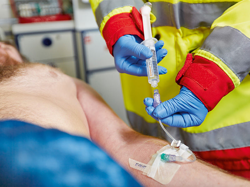 Patient covered with a blue blanket from the middle of the abdomen lies inside an ambulance. The patient's left forearm is punctured with a green Introcan Safety® 3 cannula. Caresite valve with infusion line and white sliding clamp is connected. Fixation with cannula plaster. Person in emergency jacket and blue gloves holds filled syringe in right hand. Left hand holds line below the valve.
