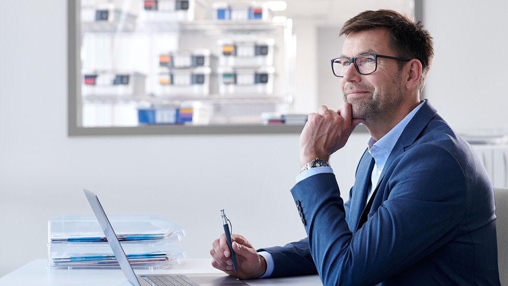 A man with glasses is sitting in a modern office, using a laptop, with a very thoughtful expression on his face