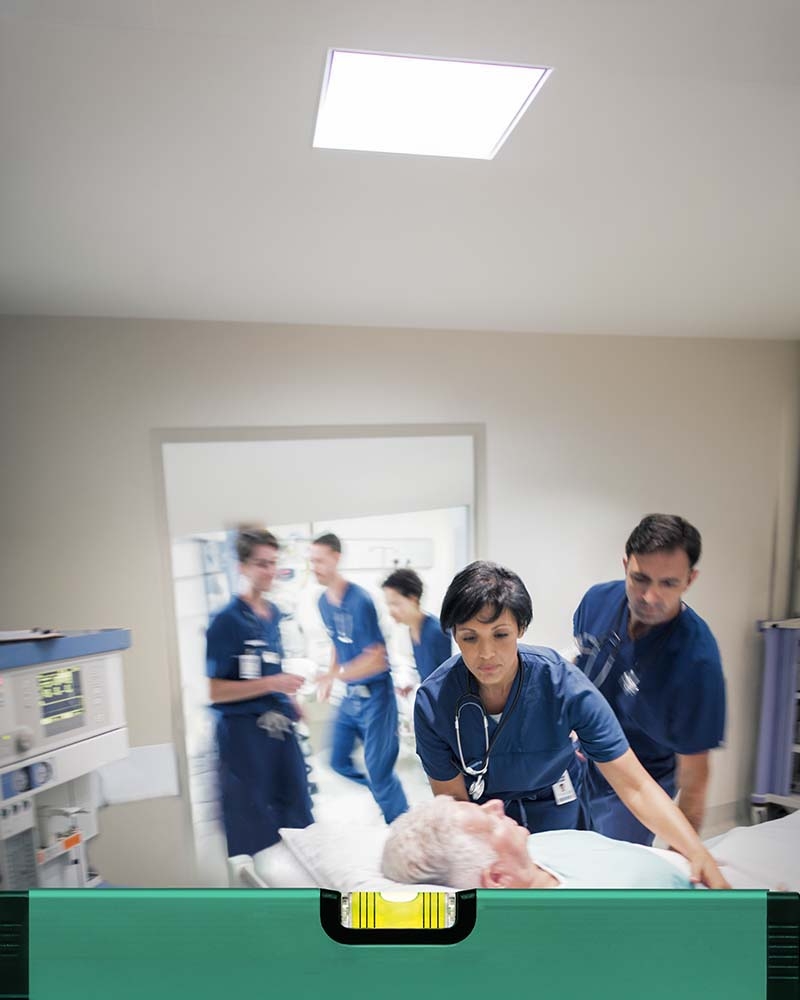 A medical team in clinical workwear is accompanying a person on a hospital bed with acute sepsis through a clinical corridor. In the foreground, a green measuring tool with an integrated spirit level is visible. In the background, medical equipment and monitoring devices are positioned along the wall. ​