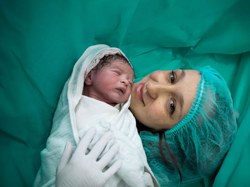 A mother lies in the operating room after a  caesarean section and looks at her newborn baby,  which a nurse is showing her. She looks exhausted,  but also relieved. She is wearing a surgical cap over  her dark hair, the newborn is wrapped in a white  cloth and is wearing a light blue cap.