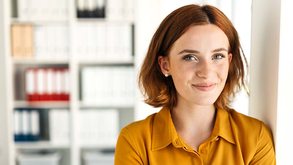 A woman is smiling confidently, looking directly into the camera, bookshelves is visible in the background.